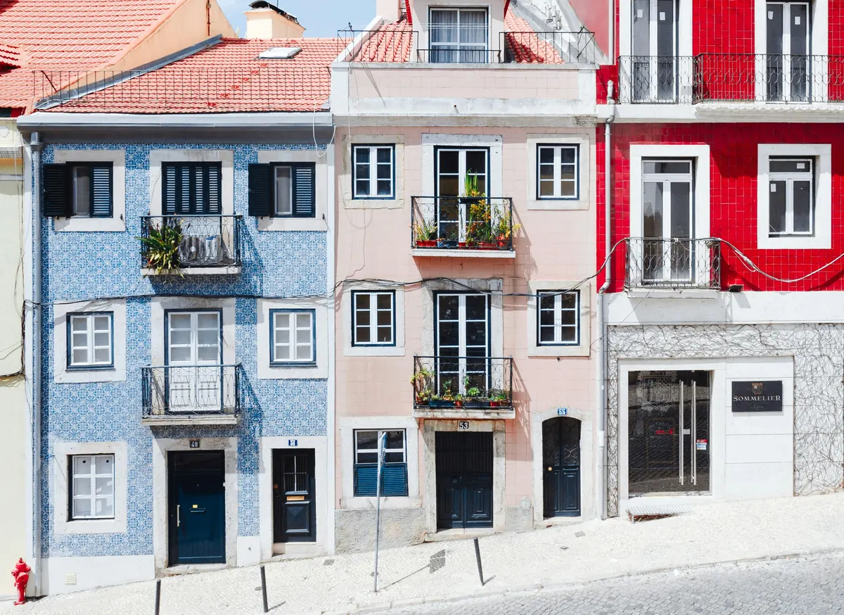 A row of Lisbon townhouses on a sloping street, one faced in blue and white azulejo tiles, one pale pink and one bright red, each with small wrought-iron balconies